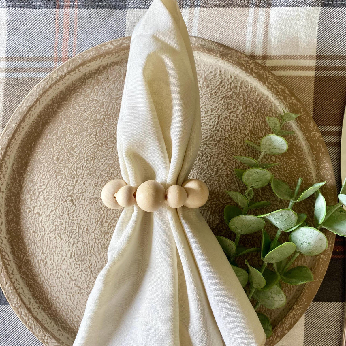 A cream-colored fabric napkin tied with a natural wood bead napkin ring, arranged on a textured beige ceramic plate with green eucalyptus leaves, set on a plaid tablecloth.