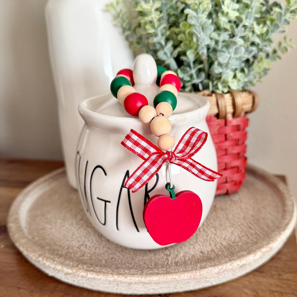 A decorative beaded sugar bowl charm with red, green, and natural wood beads, a red gingham ribbon bow, and a red apple-shaped hanging ornament, displayed on a white ceramic sugar jar.