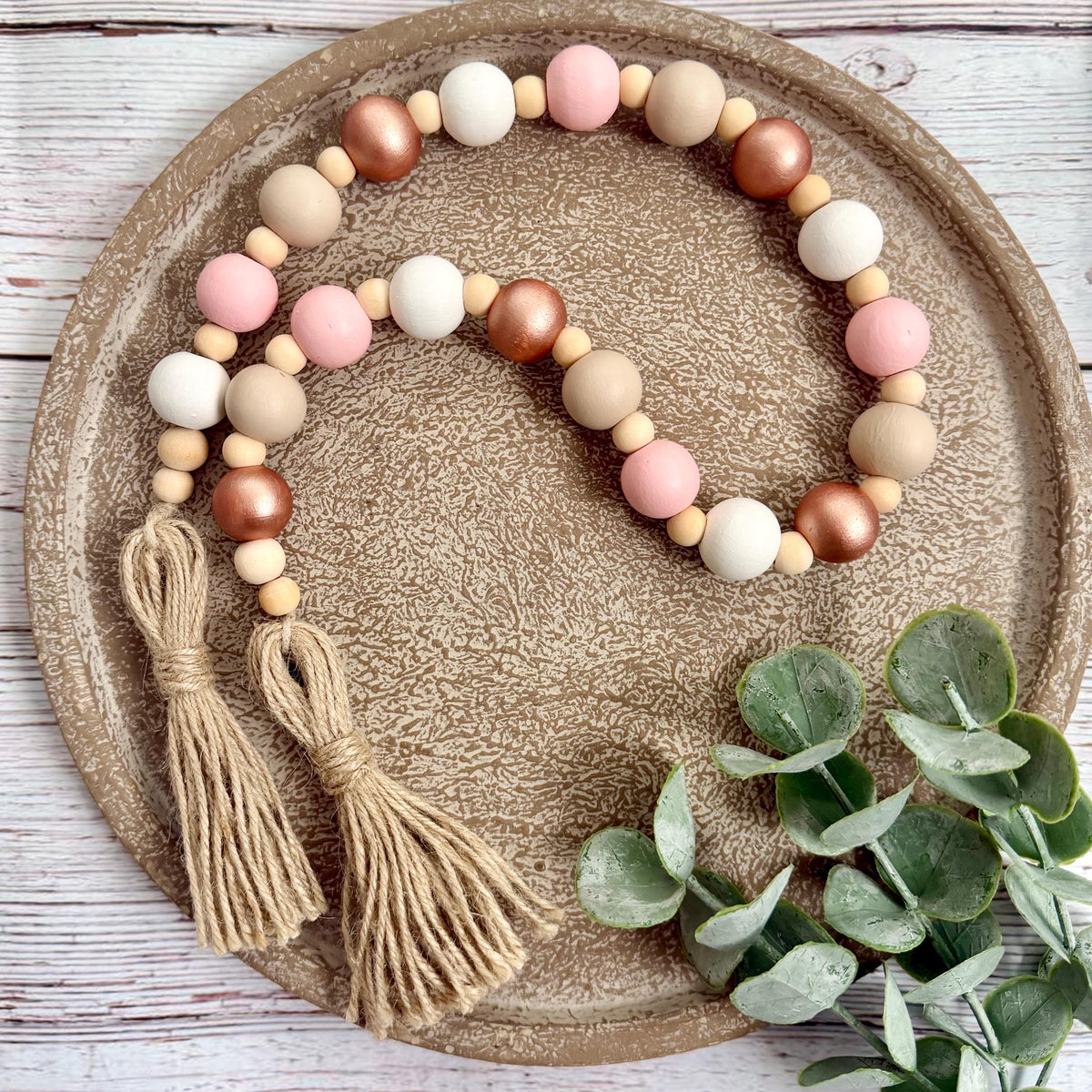 A beaded garland with two tassels, featuring matte beige, white, and pink wooden beads with metallic rose-gold beads, displayed on a textured brown ceramic plate with eucalyptus leaves.
