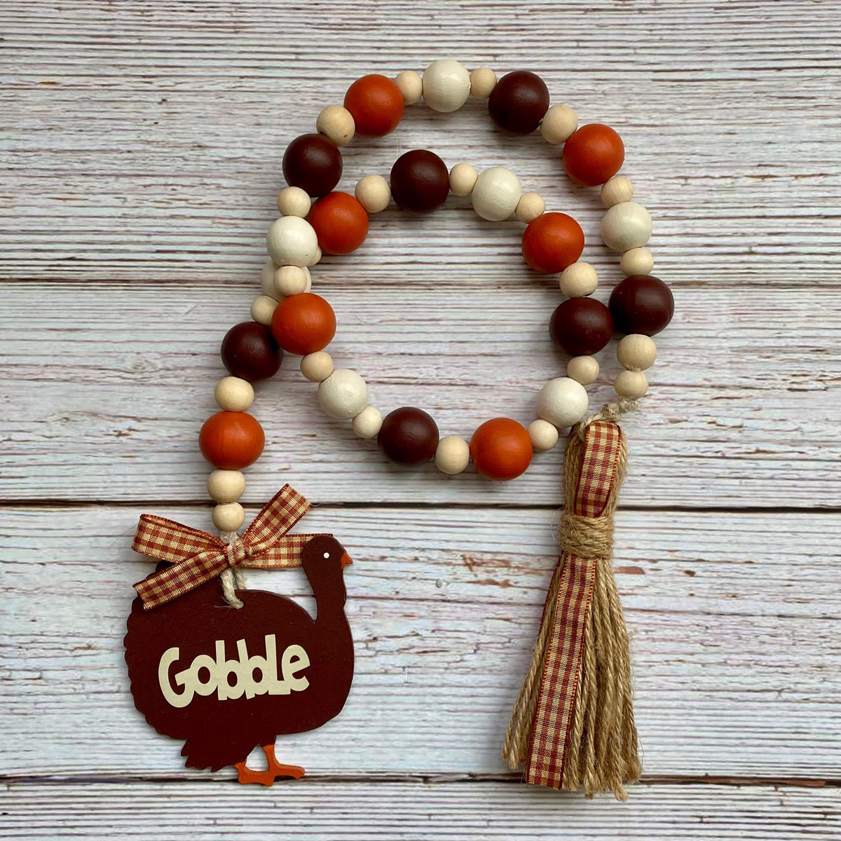 A rustic fall-themed beaded garland with orange, brown, and cream wooden beads, a dark brown turkey-shaped ornament reading 'Gobble' with a gingham bow, and a jute tassel, displayed on a whitewashed wood plank background.