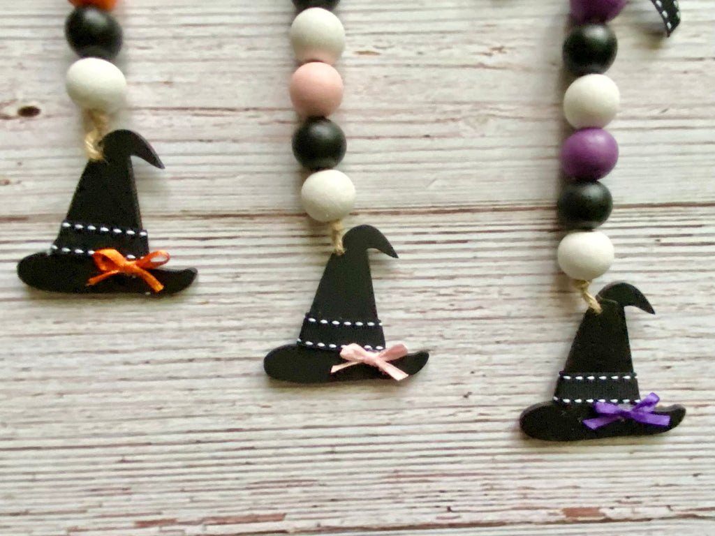 Three black wooden witch hat ornaments hanging from beaded garlands  on a whitewashed wooden surface. The necklaces feature black, white, pale pink, and purple beads, with the hats decorated with orange, pink, and purple ribbons.