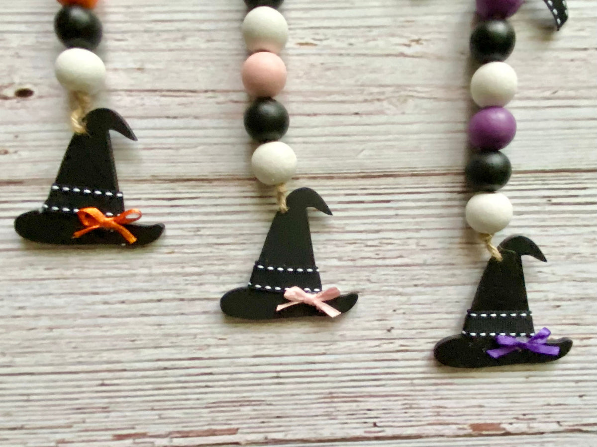 Three black wooden witch hat ornaments hanging from beaded garlands  on a whitewashed wooden surface. The necklaces feature black, white, pale pink, and purple beads, with the hats decorated with orange, pink, and purple ribbons.