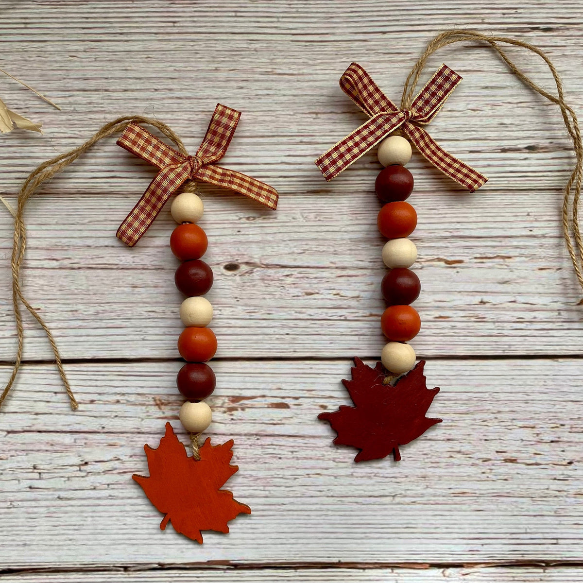 Two autumn-themed wooden bead garlands with gingham ribbon bows, red and orange maple leaf ornaments, and natural wood beads, displayed on a rustic white-washed wooden background.
