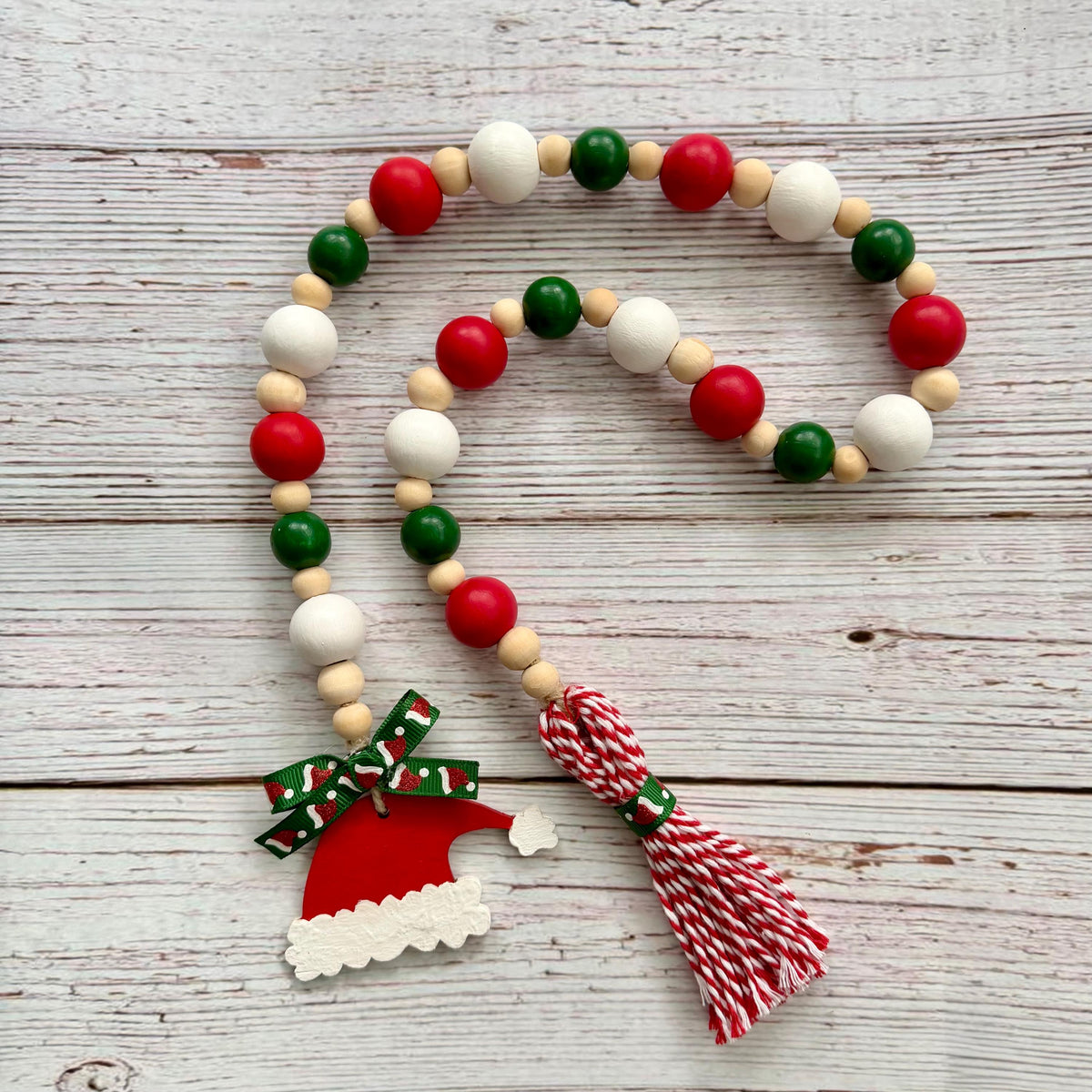 A wooden bead garland with red, green, and white beads, a Santa hat charm, and a red and white striped tassel, displayed on a rustic white-washed wooden background.