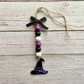 A Halloween-themed wooden bead ornament with a black witch hat, purple and white beads, a black ribbon bow, and a jute hanging string, displayed on a rustic white-washed wood background.