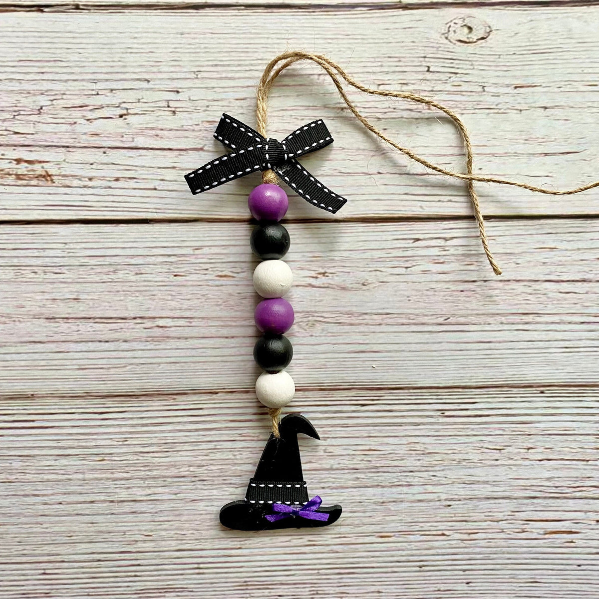 A Halloween-themed wooden bead ornament with a black witch hat, purple and white beads, a black ribbon bow, and a jute hanging string, displayed on a rustic white-washed wood background.