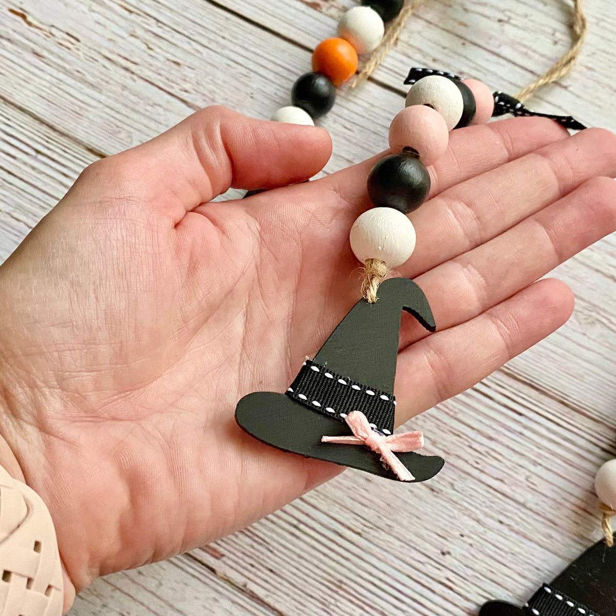 A hand holding a Halloween-themed beaded garland with wooden witch hats, black and white beads, and small orange and purple accents, set against a rustic white wood background.