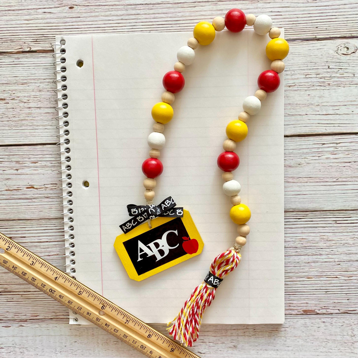 A wooden bead garland with red, yellow, and natural wood beads, featuring a yellow and black 'ABC Kindergarten' chalkboard charm and a red, yellow, and white tassel, displayed on a lined notebook with a wooden ruler.