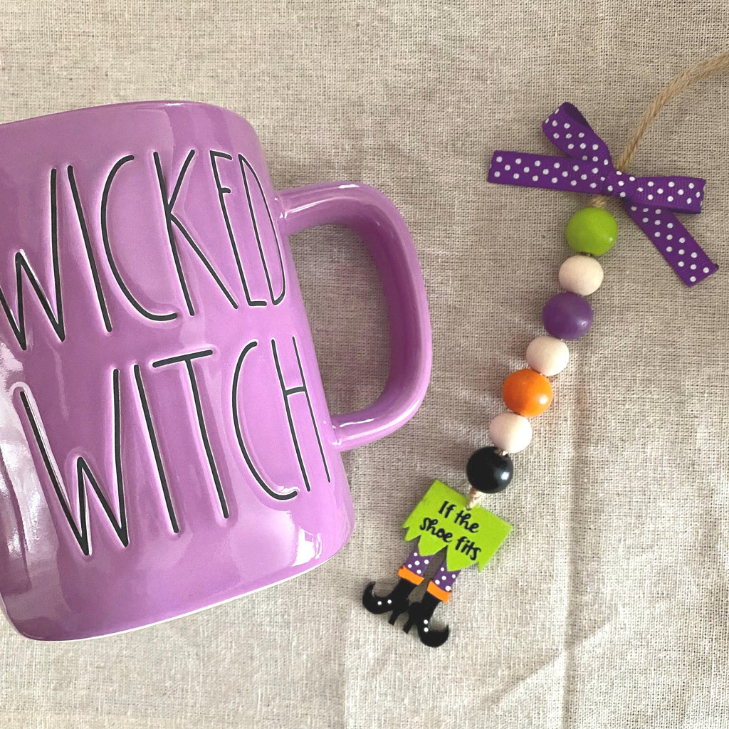 A purple ceramic mug with 'WICKED WITCH' printed on it in black, shown next to a beaded hanging ornament with a purple polka-dot bow and small witch-themed charms on a beige fabric background.