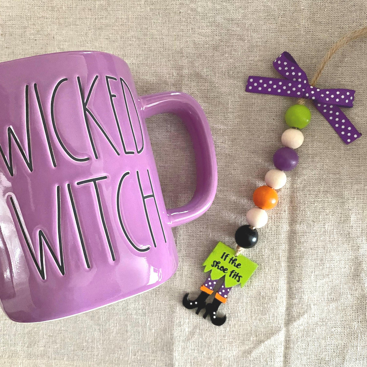 A purple ceramic mug with 'WICKED WITCH' printed on it in black, shown next to a beaded hanging ornament with a purple polka-dot bow and small witch-themed charms on a beige fabric background.