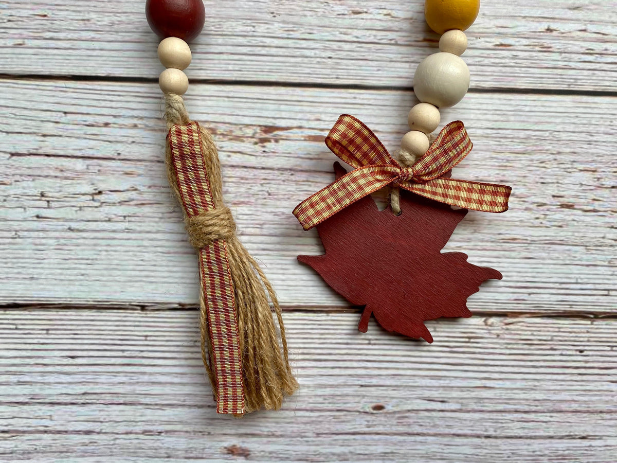 Autumn-themed wooden bead garland with a tassel and maple leaf ornament on a whitewashed wooden background.