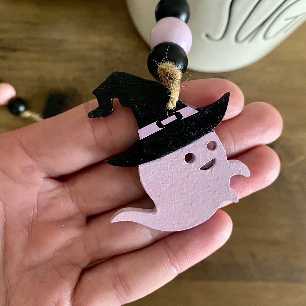 A hand holding a small Halloween decoration featuring a light pink ghost wearing a black witch hat, with a beaded garland hanging above it on a wooden surface.