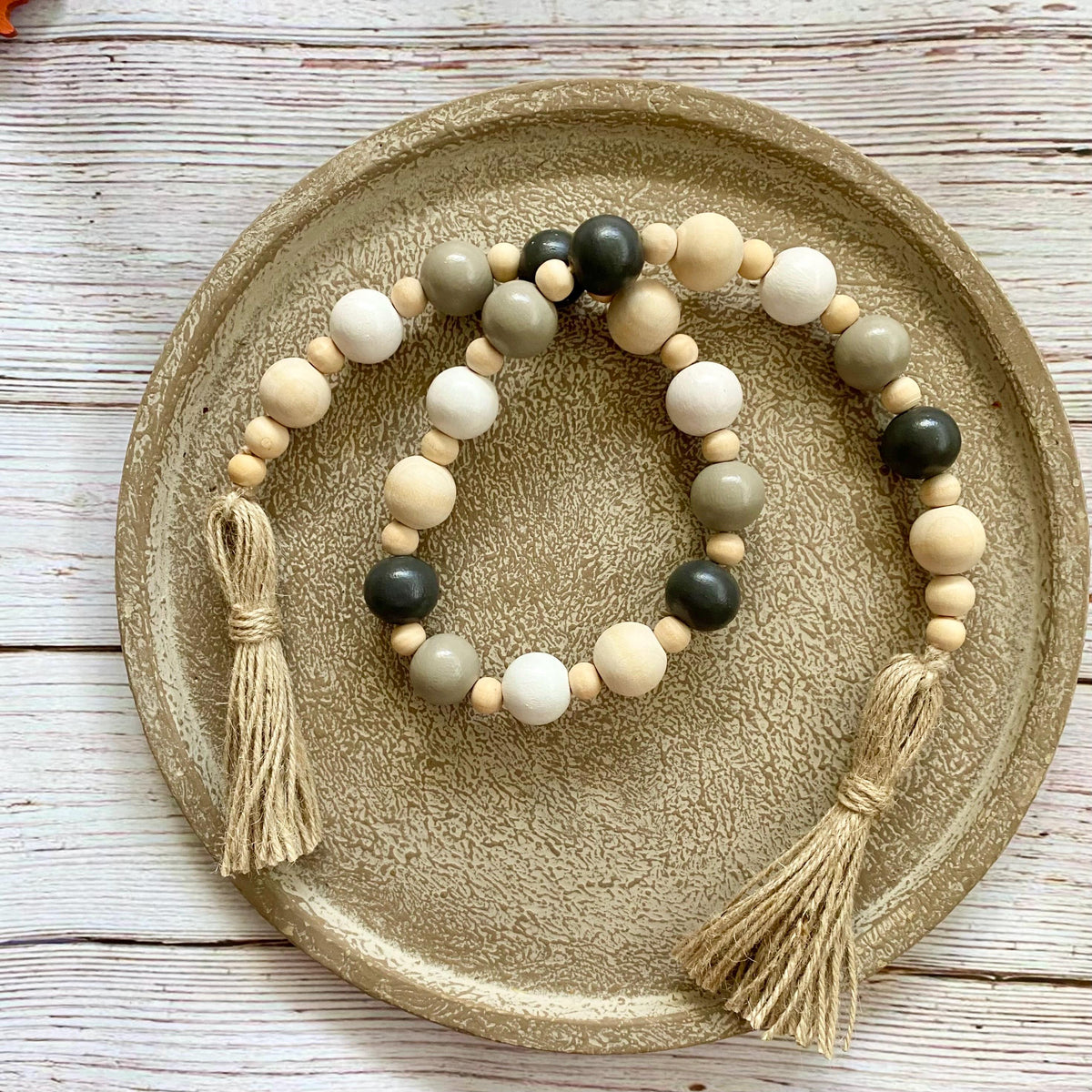 Three beaded garland with wooden beads and tassels, arranged on a textured round tray over a whitewashed wooden surface.