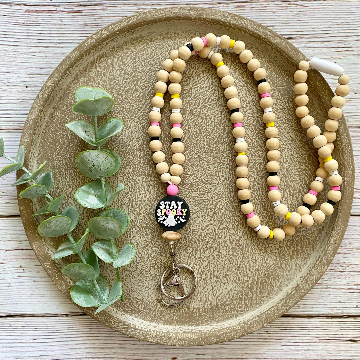 A beaded necklace with natural wood beads, pink, yellow, and black accent beads, and a black round charm that reads 'STAY spooky' in white text, displayed on a round textured tray with eucalyptus leaves.