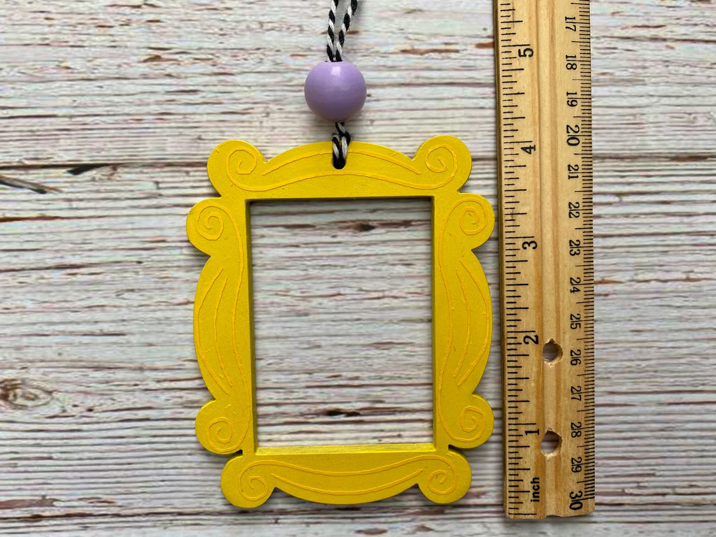 A yellow ornate picture frame ornament with a lavender bead and black-and-white string, shown next to a wooden ruler on a whitewashed wood background.