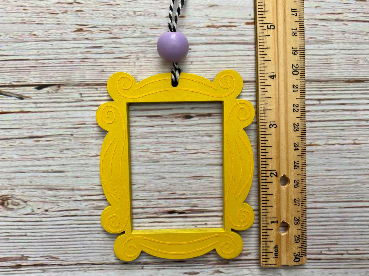 A yellow ornate picture frame ornament with a lavender bead and black-and-white string, shown next to a wooden ruler on a whitewashed wood background.