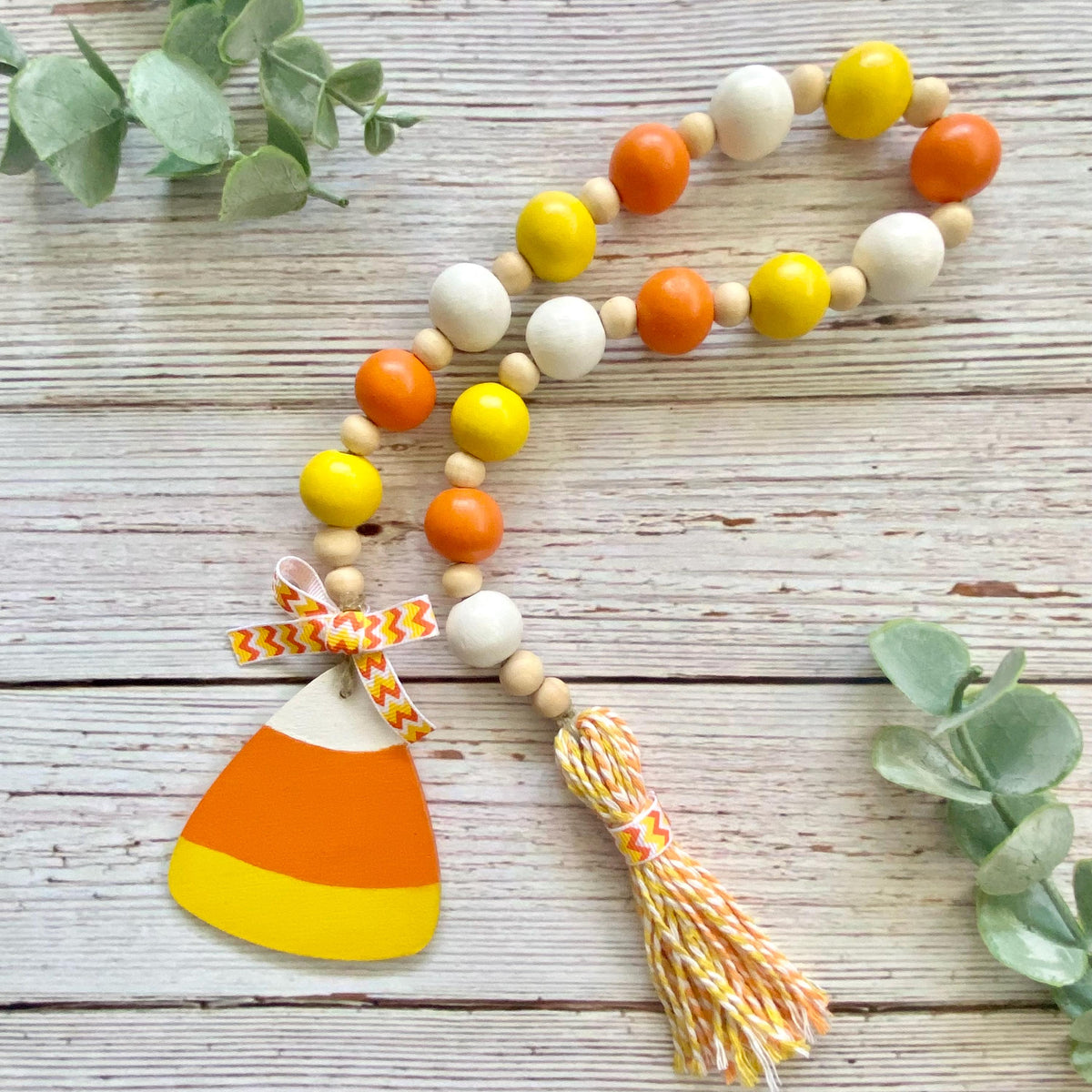 A wooden bead garland with orange, yellow, and white beads, featuring a hanging wooden candy corn ornament tied with a matching striped ribbon and a yellow, orange, and white tassel, displayed on a rustic white wood background with green eucalyptus leaves.