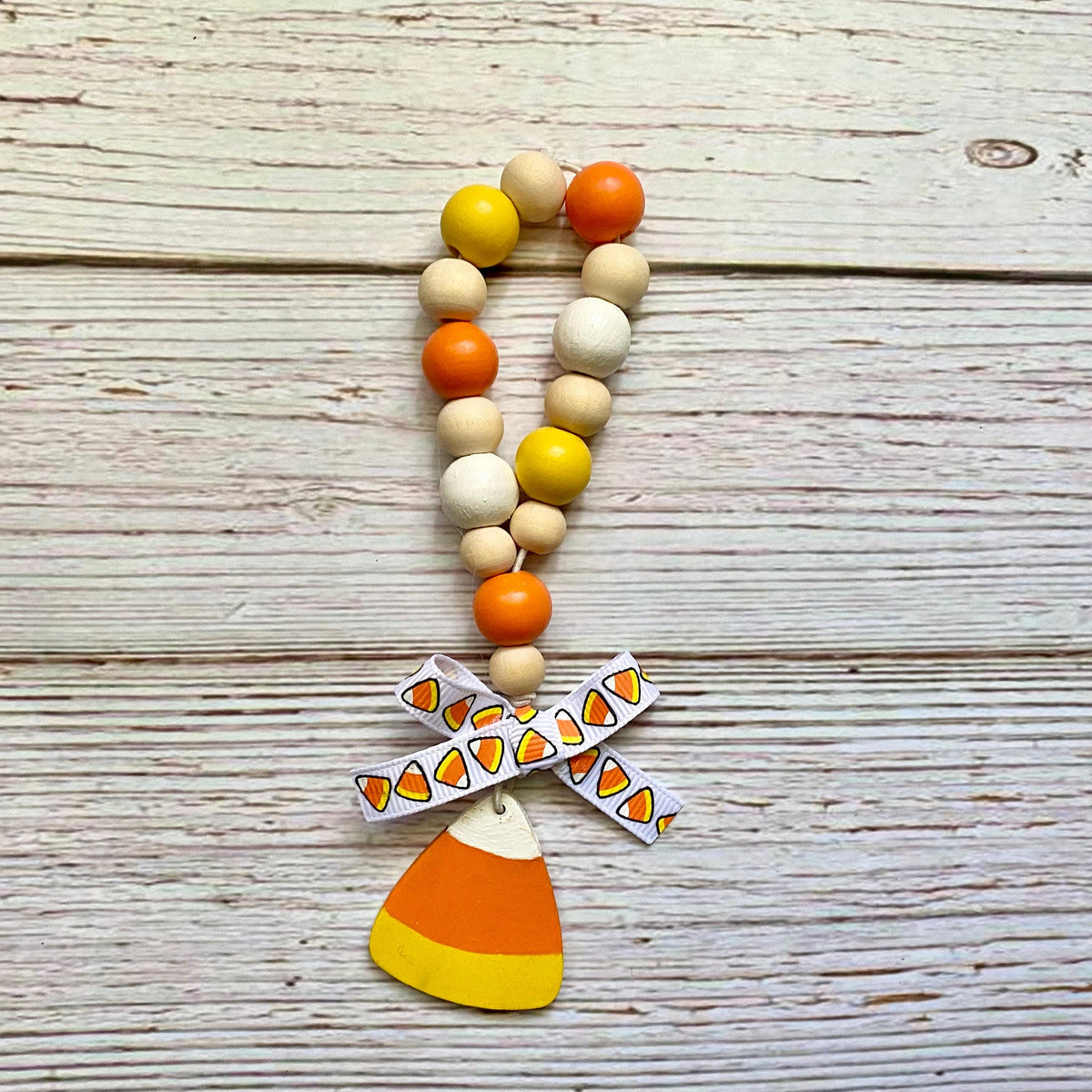 A beaded loop with orange, yellow, and natural wood beads, featuring a white ribbon bow printed with candy corn motifs and a wooden candy corn charm, displayed on a light wood plank background.