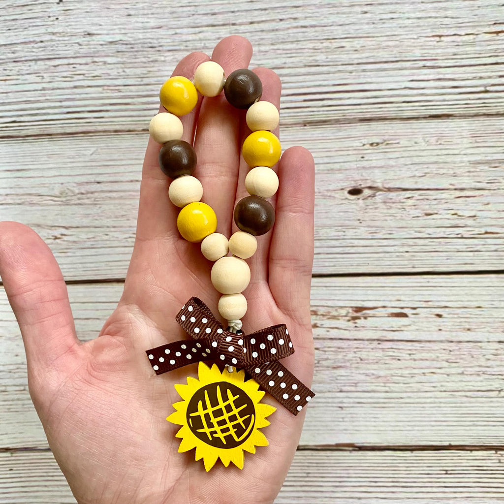 A hand holding a beaded charm bracelet with yellow, brown, and cream wooden beads, a brown polka dot ribbon bow, and a yellow sunflower charm, set against a rustic white wood plank background.