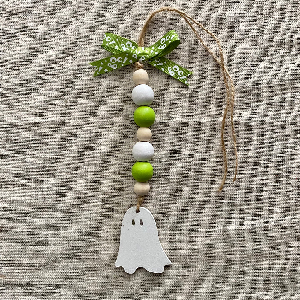 A handmade wooden bead hanging ornament with a white ghost cutout, green and beige wooden beads, a green patterned ribbon, and jute twine, displayed on a textured beige linen background.