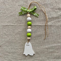 A handmade wooden bead hanging ornament with a white ghost cutout, green and beige wooden beads, a green patterned ribbon, and jute twine, displayed on a textured beige linen background.