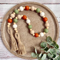 A beaded garland with natural wood beads, orange and green round beads, and two jute tassels, arranged on a textured round tray with eucalyptus leaves.