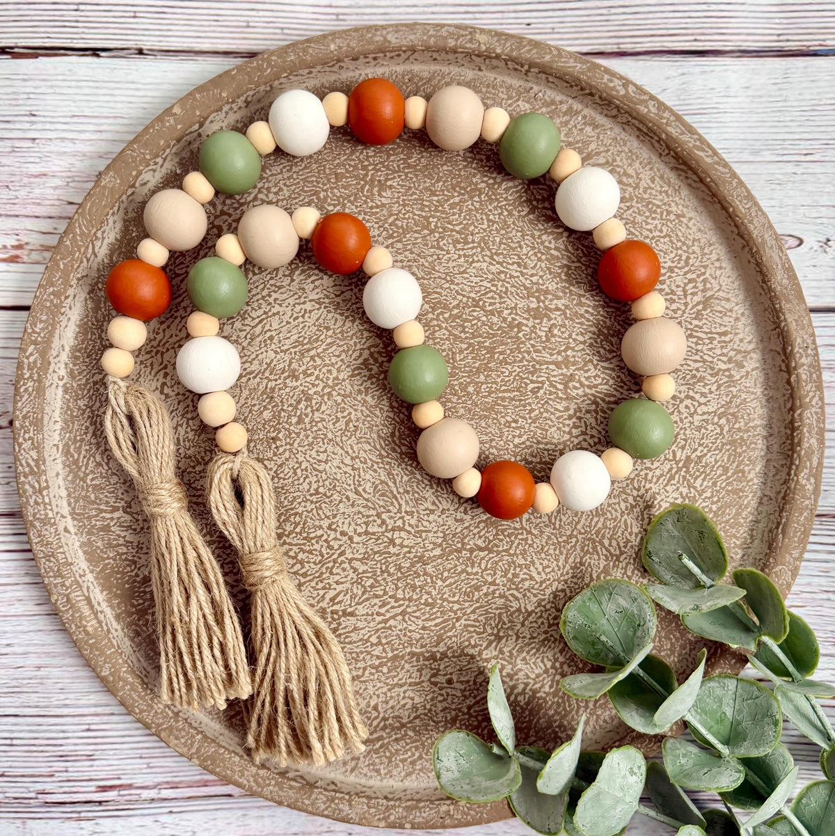 A beaded garland with natural wood beads, orange and green round beads, and two jute tassels, arranged on a textured round tray with eucalyptus leaves.