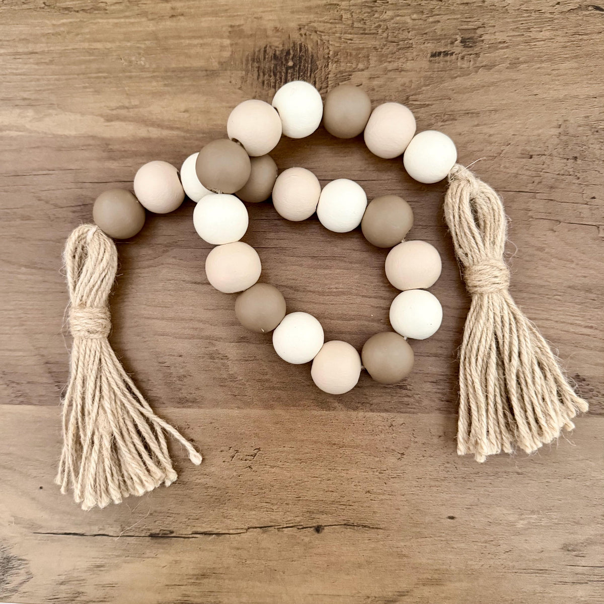 A bohemian-style wooden bead garland with two knotted jute tassels, displayed on a rustic wooden surface. The beads are in alternating shades of natural wood brown, cream, and off-white.