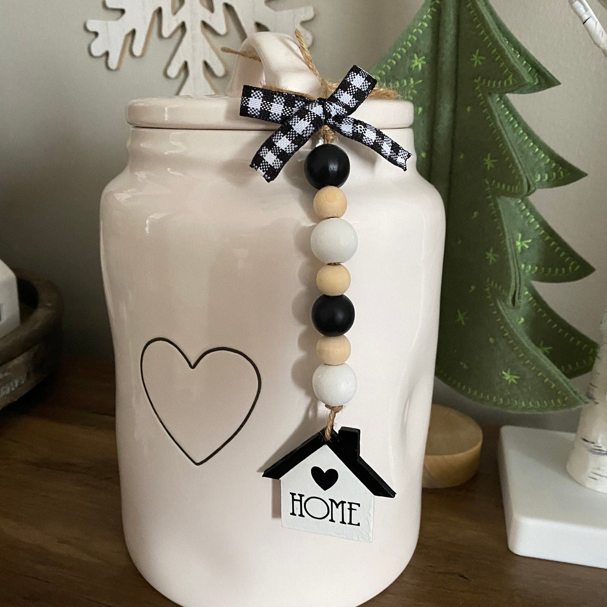 A white ceramic jar with a lid, decorated with a black heart outline, a beaded garland with a black-and-white gingham bow, and a small house-shaped tag that says HOME, displayed on a wooden surface with holiday decor in the background.