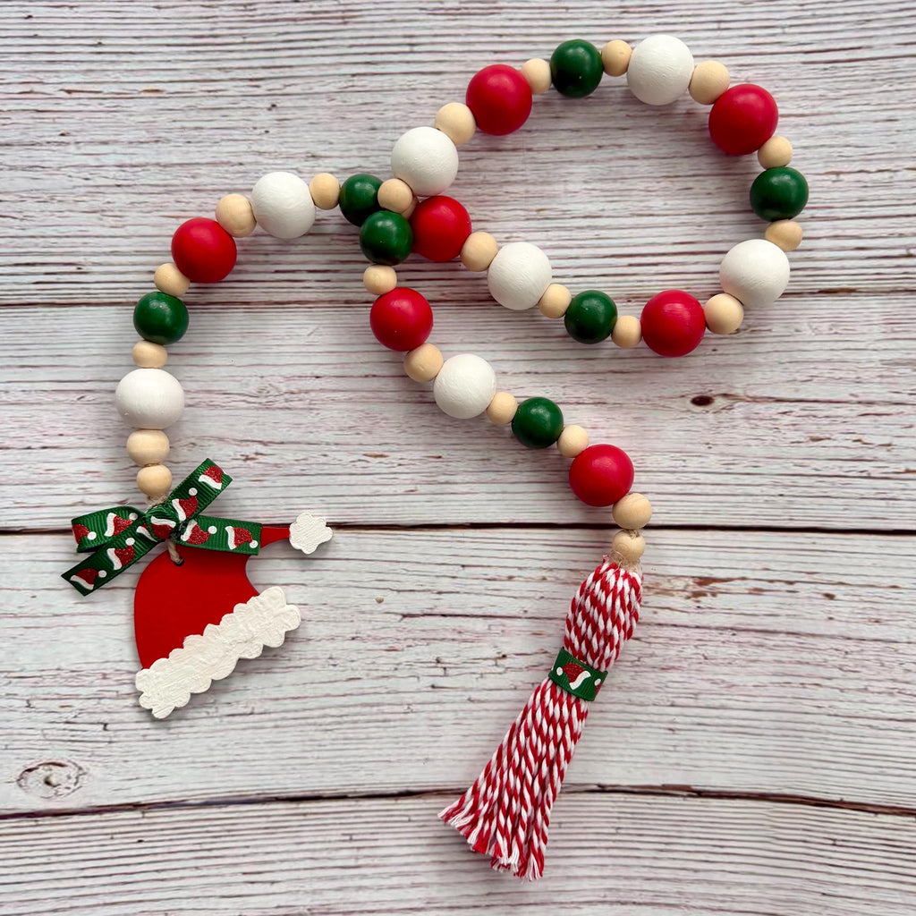 A Christmas-themed wooden bead garland with red, green, and white wooden beads, a Santa hat charm, a red and white patterned bow, and a red and white striped tassel, displayed on a light gray wooden plank background.