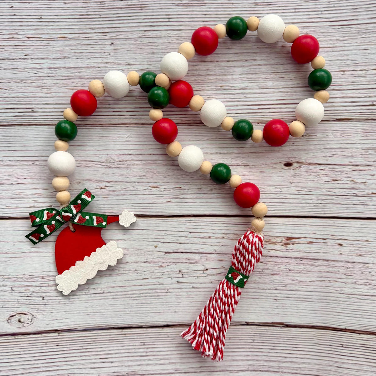 A Christmas-themed wooden bead garland with red, green, and white wooden beads, a Santa hat charm, a red and white patterned bow, and a red and white striped tassel, displayed on a light gray wooden plank background.