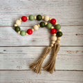 A wooden bead garland with alternating red, sage green, olive green, and natural beige beads, finished with two knotted jute tassels, displayed on a rustic white-washed wooden background.