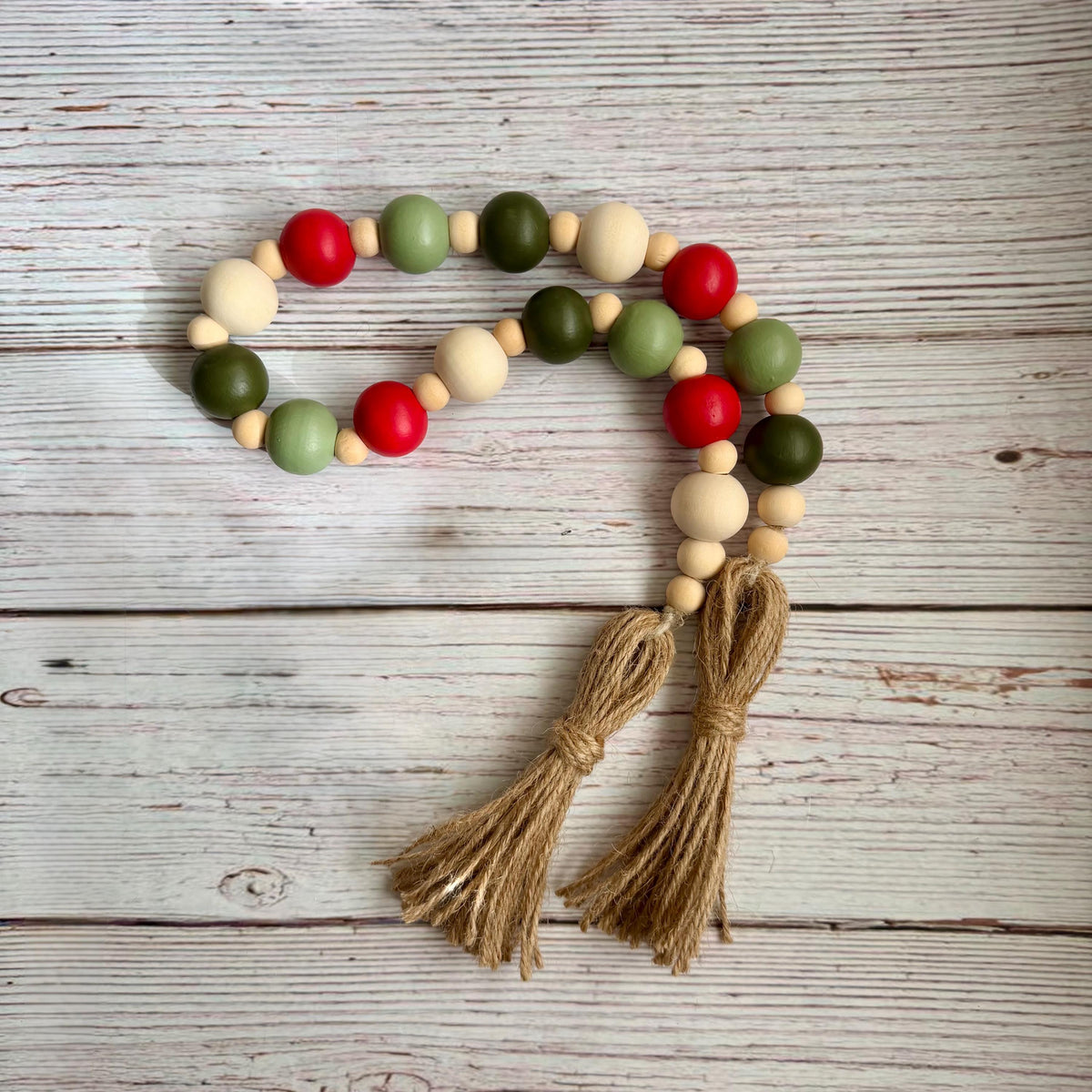 A wooden bead garland with alternating red, sage green, olive green, and natural beige beads, finished with two knotted jute tassels, displayed on a rustic white-washed wooden background.