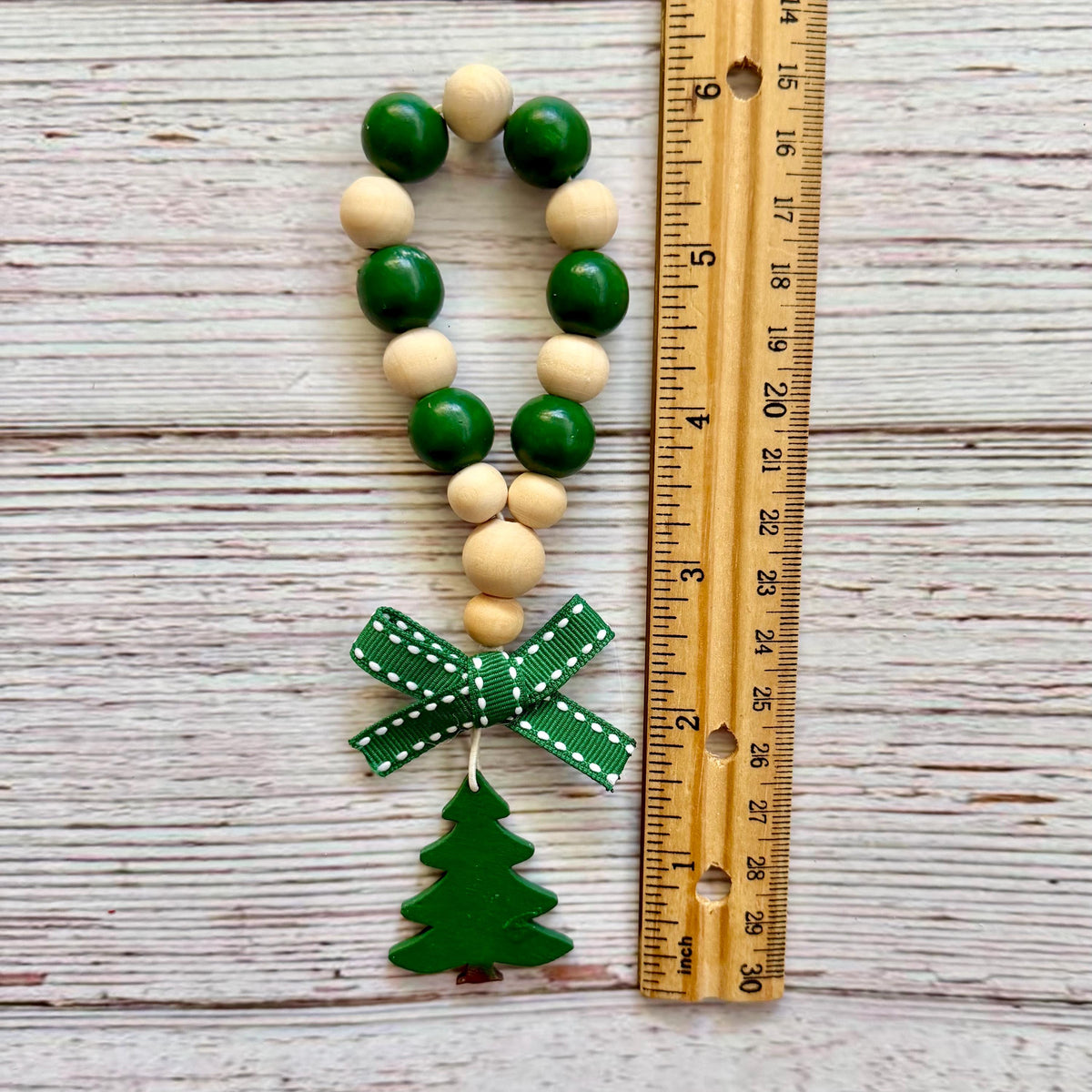 A beaded garland ornament with green and natural wood beads, a green ribbon bow, and a green Christmas tree charm, shown next to a wooden ruler on a whitewashed wood background.