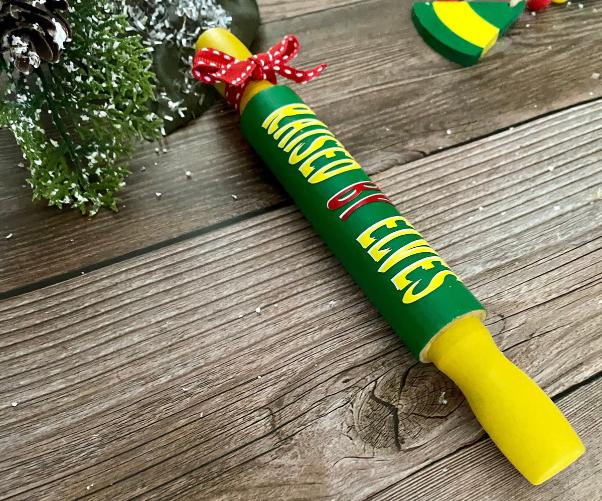 A Christmas-themed wooden rolling pin ornament with 'Kissed by Elves' written in yellow and red lettering on a green body, featuring a red and white polka dot bow at the top and a yellow handle, displayed on a rustic wooden surface.