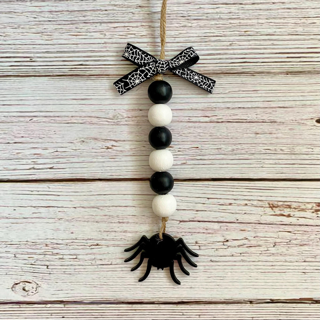A hanging Halloween ornament made from a jute rope, featuring a black and white spiderweb-patterned bow, alternating black and white wooden beads, and a black spider at the bottom, displayed against a rustic whitewashed wood plank background.
