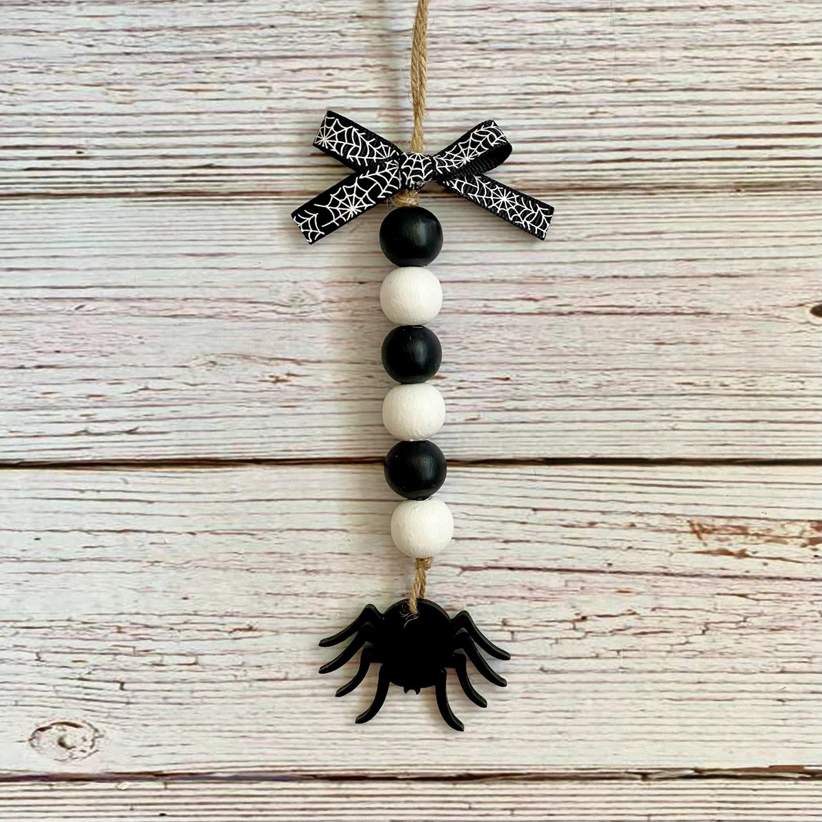 A hanging Halloween ornament made from a jute rope, featuring a black and white spiderweb-patterned bow, alternating black and white wooden beads, and a black spider at the bottom, displayed against a rustic whitewashed wood plank background.