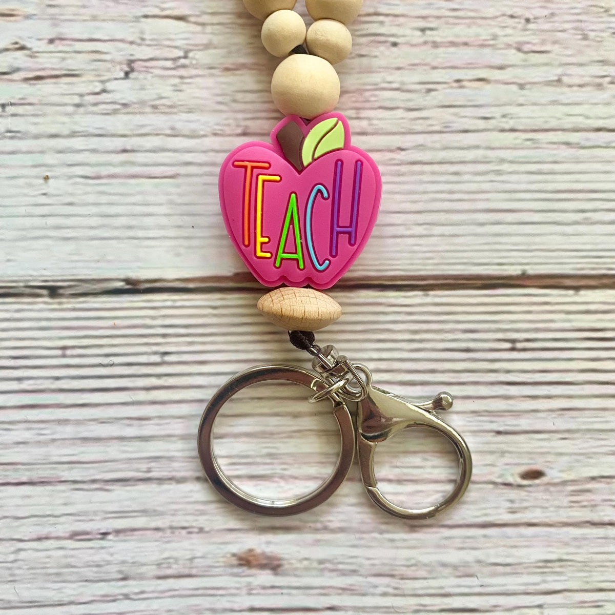 A beaded keychain with a pink heart-shaped charm spelling 'TEACH' in colorful letters, hanging from natural wood beads and attached to a silver-tone split ring on a whitewashed wood background.