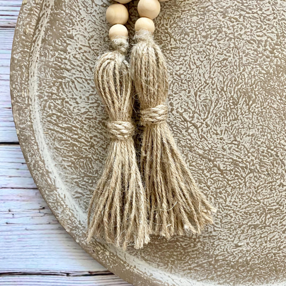 Two natural jute tassels with wooden beads, displayed on a textured beige ceramic plate over a white wooden surface.