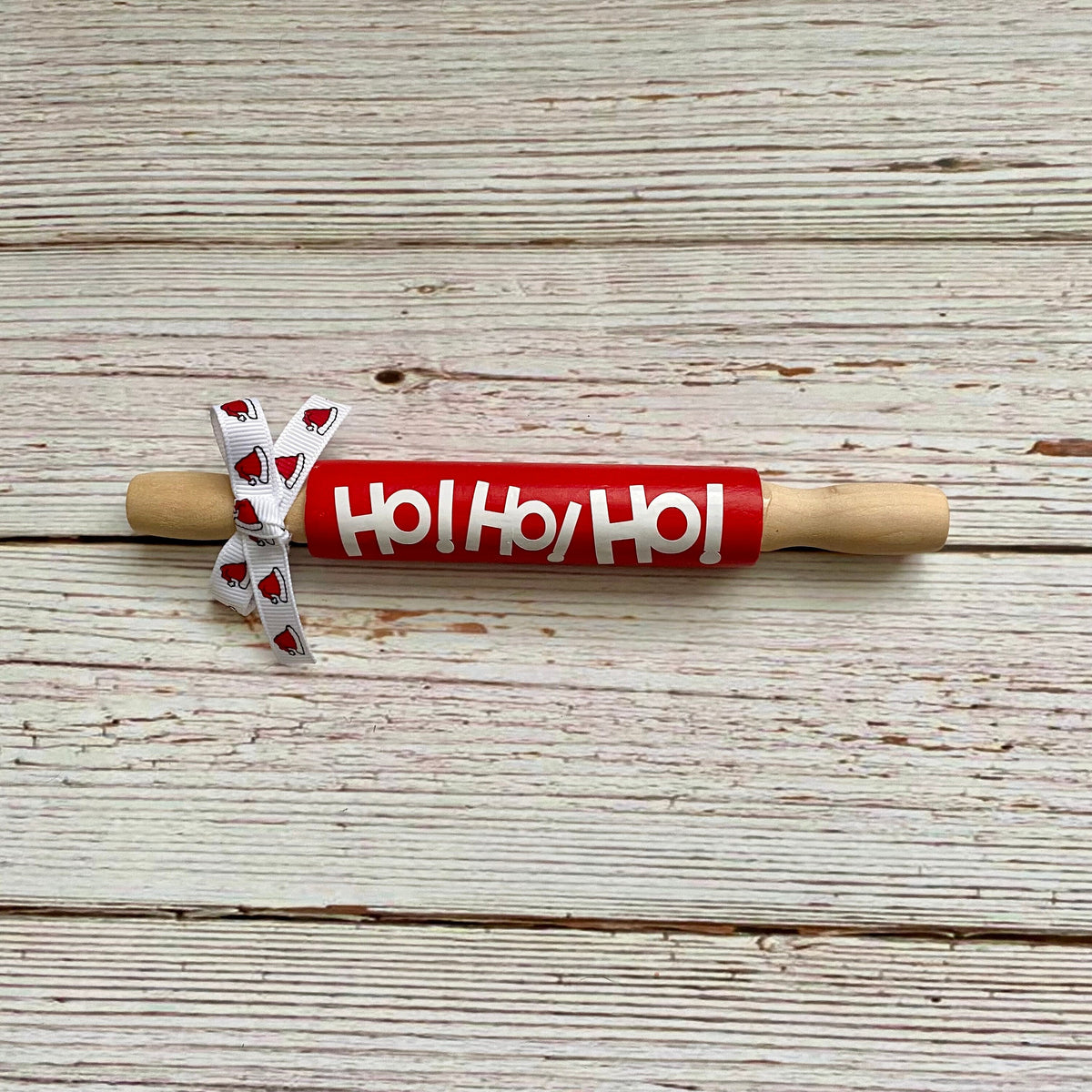 A wooden rolling pin with a bright red painted barrel printed with 'Ho Ho Ho' in white, resting on a distressed white wood plank surface. A white ribbon with red hearts is tied to the handle.