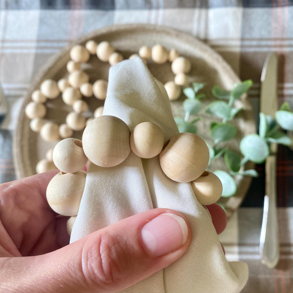 A hand holding a cream-colored fabric napkin wrapped with a string of light natural wood beads, set against a rustic table setting with a plaid tablecloth, a woven plate, and greenery.