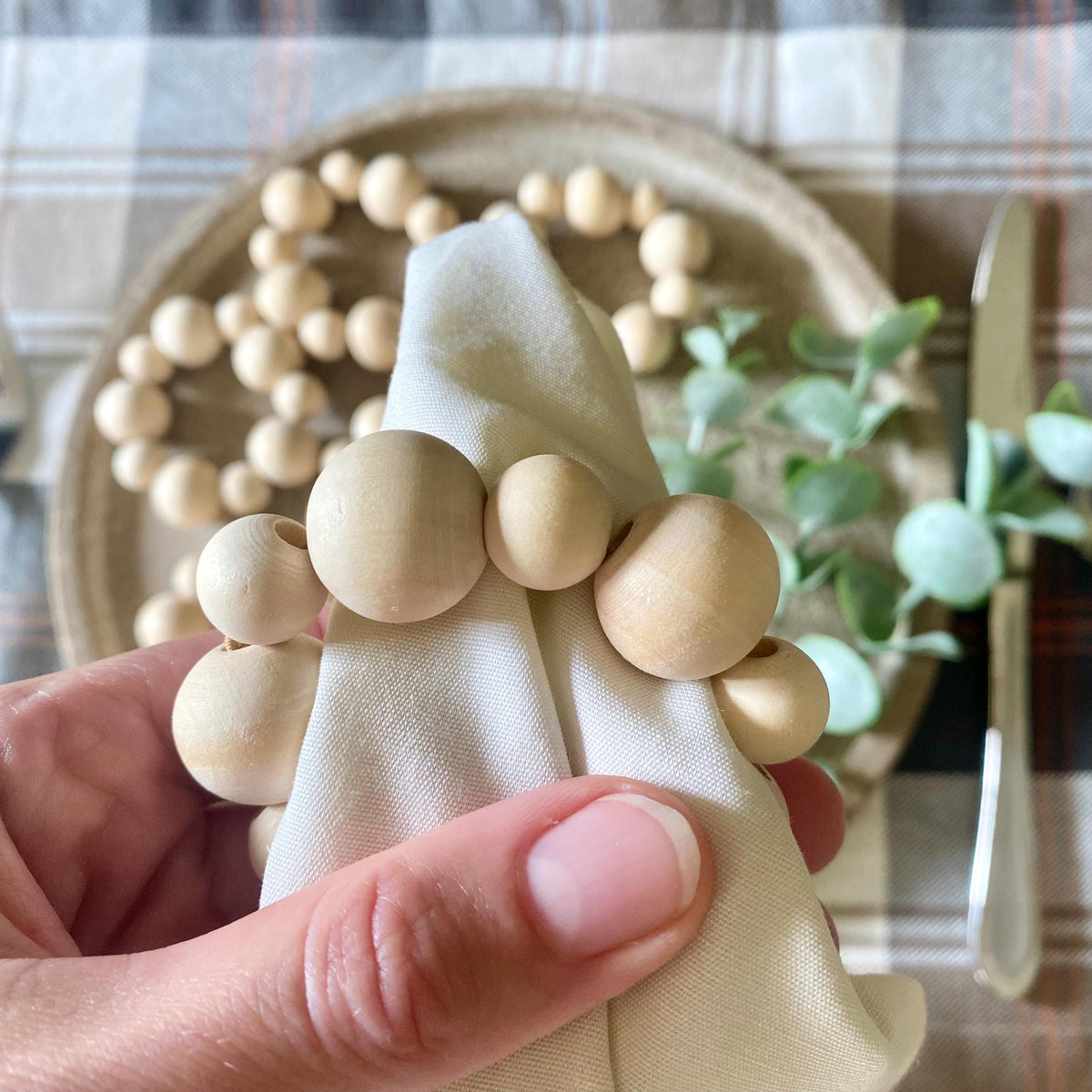 A hand holding a cream-colored fabric napkin wrapped with a string of light natural wood beads, set against a rustic table setting with a plaid tablecloth, a woven plate, and greenery.