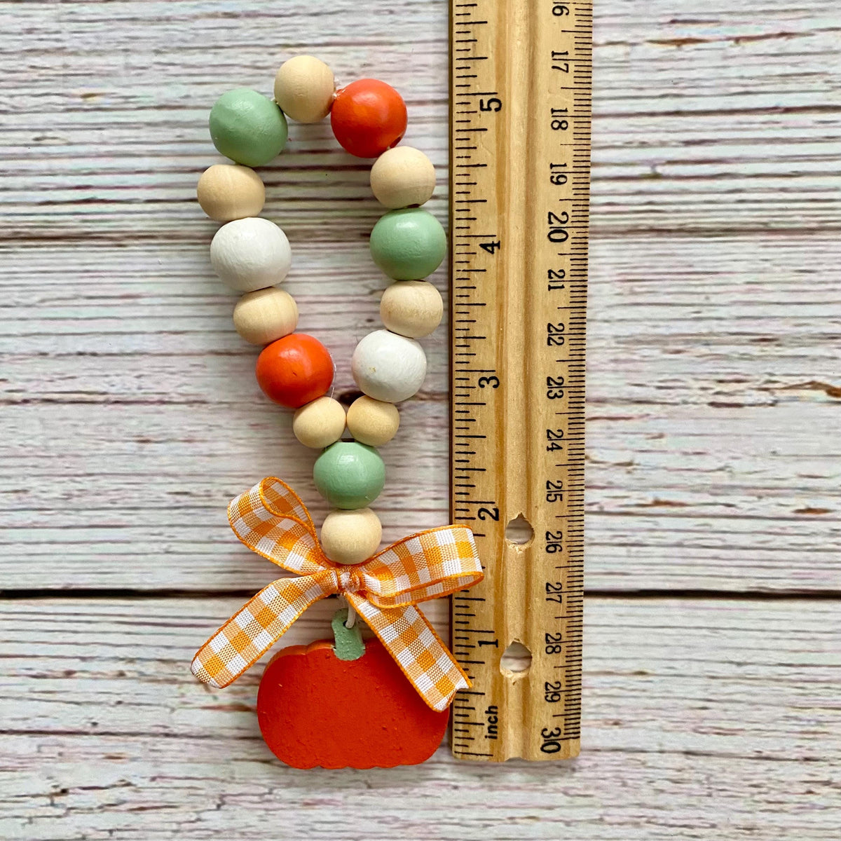A wooden bead loop with orange, mint green, and cream beads, finished with a gingham ribbon and a painted orange pumpkin charm, shown next to a wooden ruler on a whitewashed wood background.