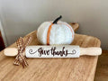 A white wooden rolling pin with 'Give thanks' written on it in black script, styled on a wooden board next to a small white knit pumpkin with orange stripes and a brown plaid ribbon.