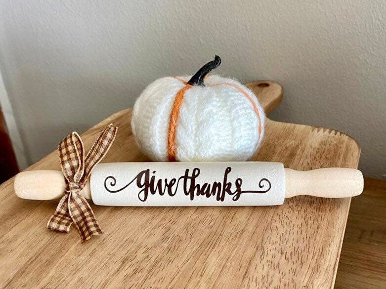 A white wooden rolling pin with 'Give thanks' written on it in black script, styled on a wooden board next to a small white knit pumpkin with orange stripes and a brown plaid ribbon.