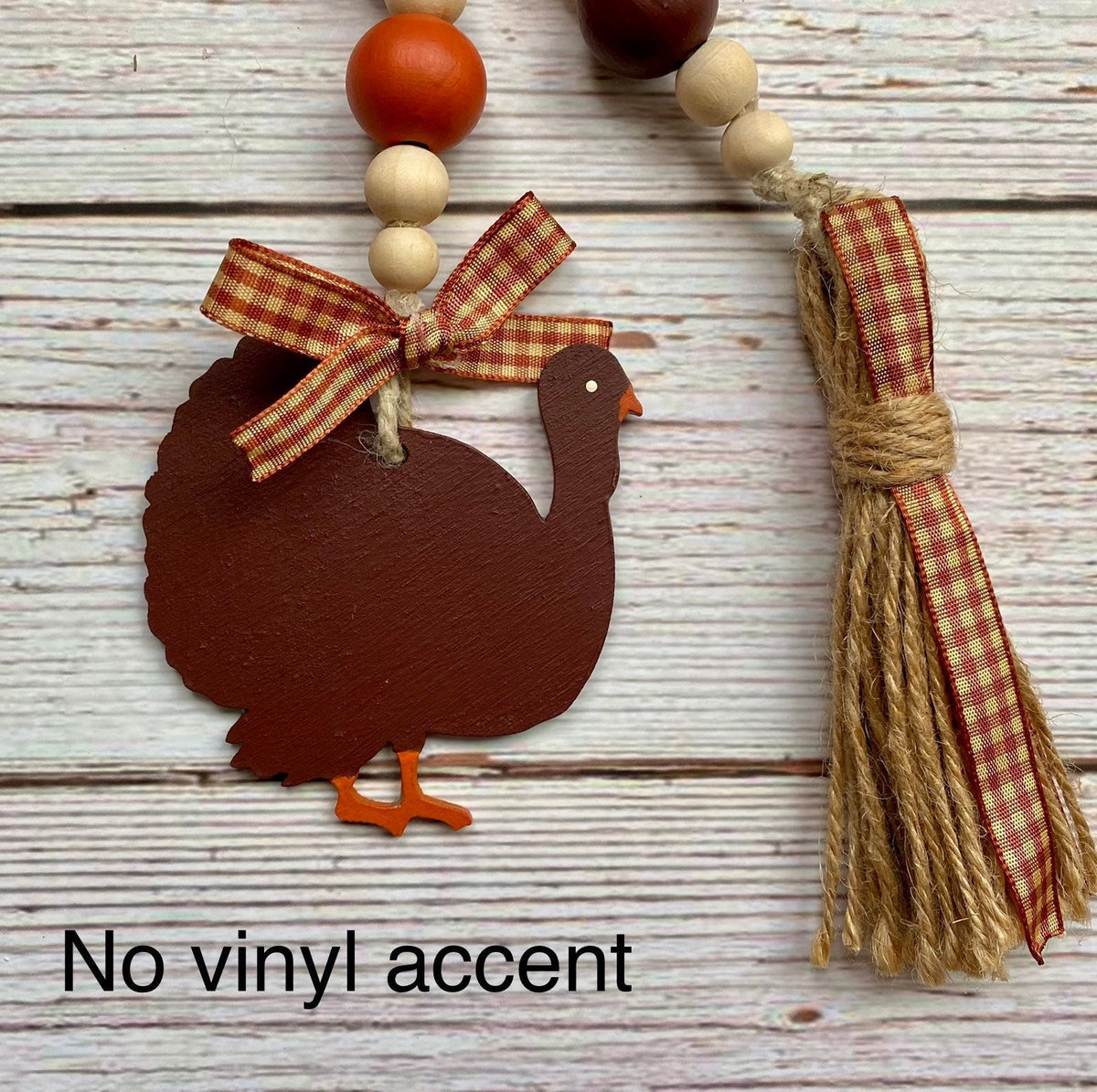 A rustic fall-themed bead garland featuring a dark brown wooden turkey ornament with a red gingham ribbon bow, orange feet, and a matching tassel on a whitewashed wood plank background.