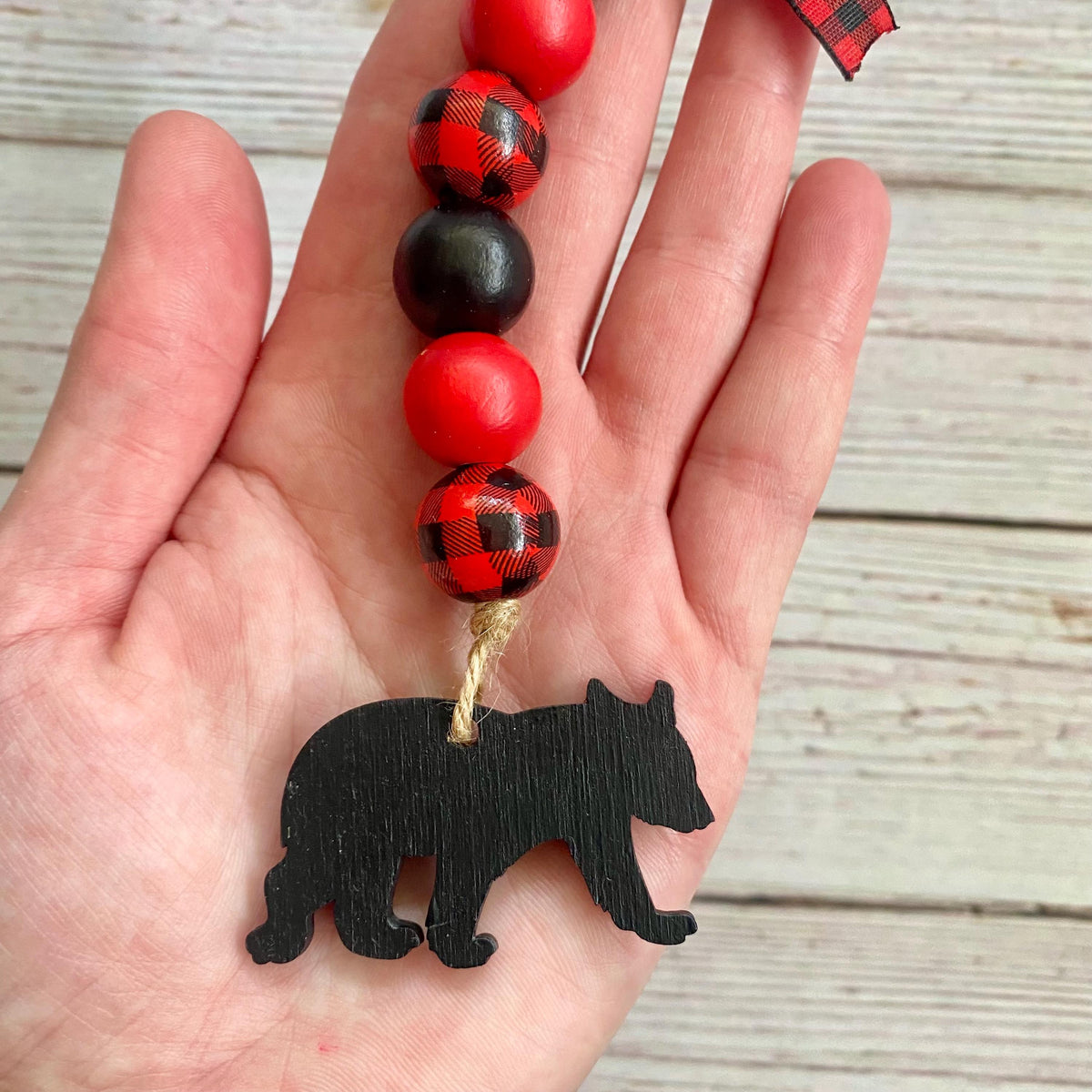 A hand holding a beaded garland with red and black buffalo plaid beads, solid red beads, and a black wooden bear silhouette charm, set against a light wood plank background.