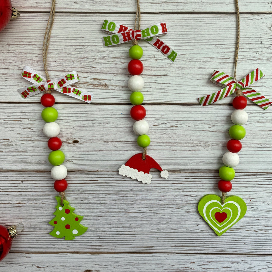 Three festive Christmas bead garlands with red, green, and white beads, hanging on a whitewashed wooden surface. The left ornament has a green Christmas tree, the middle has a Santa hat, and the right has a green and red heart.