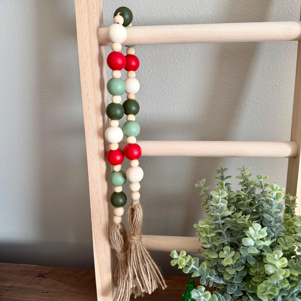 A wooden bead garland with red, green, and white beads hanging from a light wood ladder, featuring a natural jute tassel at the bottom. A small green plant is visible in the lower right corner against a neutral gray wall.