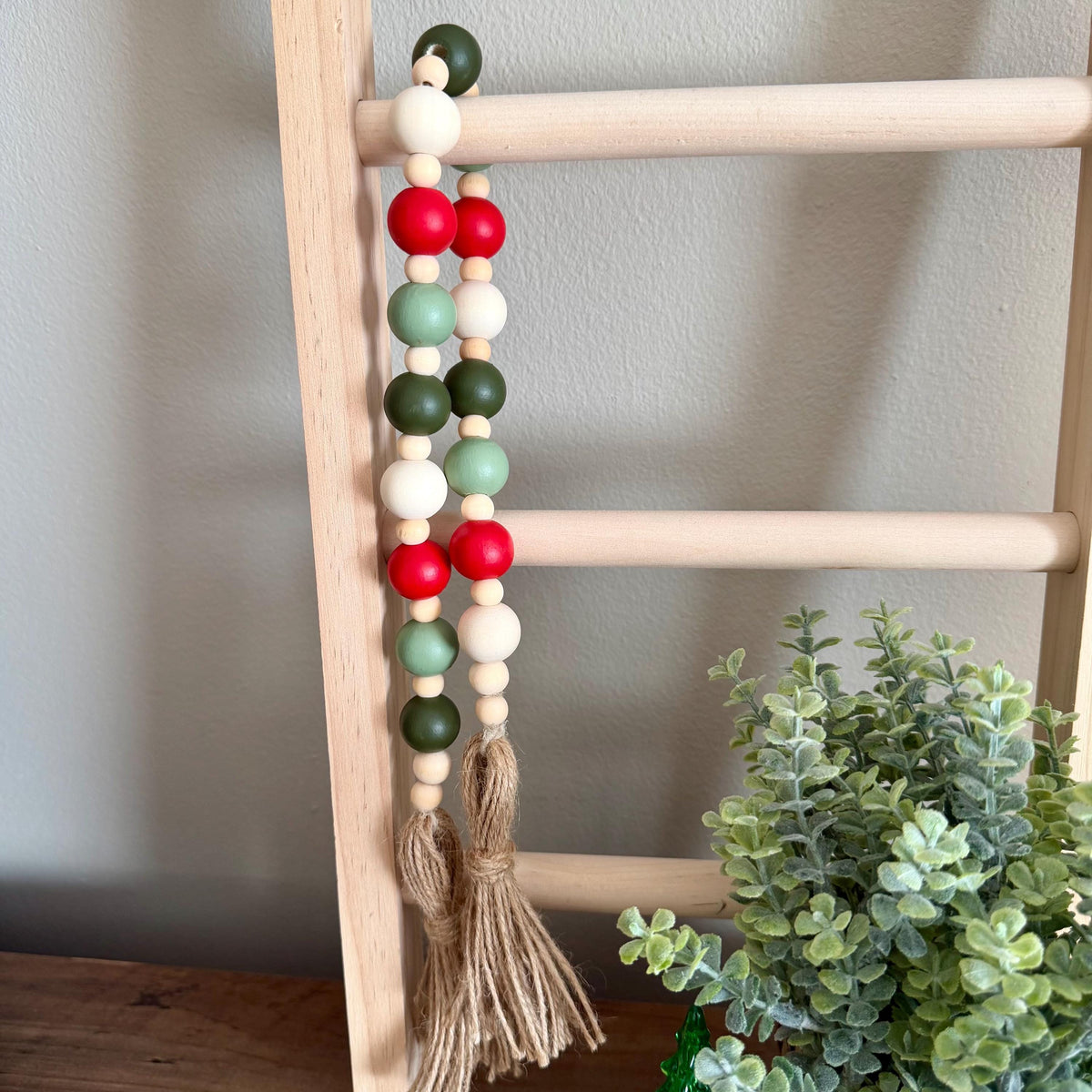A wooden bead garland with red, green, and white beads hanging from a light wood ladder, featuring a natural jute tassel at the bottom. A small green plant is visible in the lower right corner against a neutral gray wall.